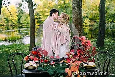 Newlyweds kissing under the plaid next to the festive table. Bride and groom in the park. Autumn wedding. Artwork Stock Photo