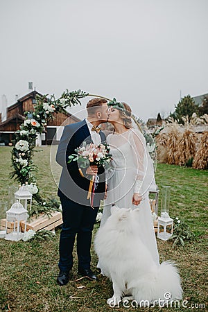 Newlyweds stand near wedding arch and kiss next to dog Stock Photo