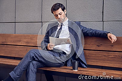 Daily news. Full length of a pleasant smiling businessman reading a newspaper with coffee while sitting on the bench Stock Photo