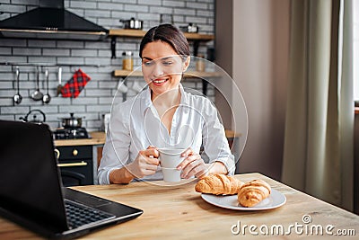 Nice cheerful businesswoman sit at table in kitchen and look at laptop. She works at home. Model hold white cup with Stock Photo