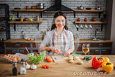 Nice cheerful woman sit at table in kitchen. She cooking. Woman look on camera and smile. She cut green onion on desk Stock Photo