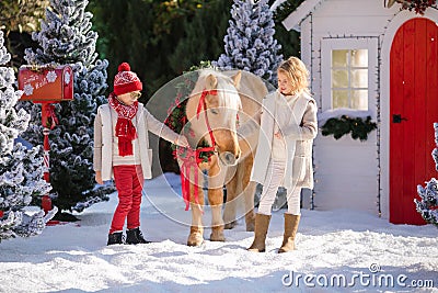 Nice children and adorable pony with festive wreath near the small wooden house and snow-covered trees. New Year and Christmas tim Stock Photo
