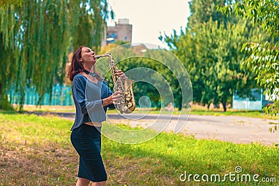 Pretty young woman with red hair in a blue sweater and black skirt plays on a yellow saxophone in a city green park Stock Photo