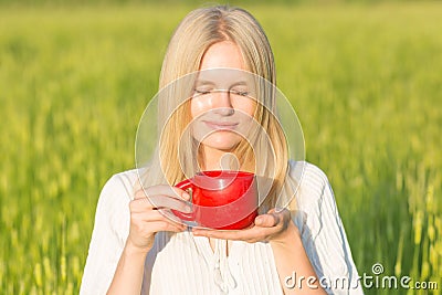 Beautiful young woman drinking tea/coffee outdoors. Green summer field background. Stock Photo