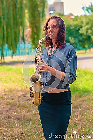 Nice girl with red hair in a blue sweater and black skirt is standing with a yellow saxophone in the forest Stock Photo