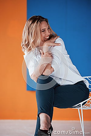 Nice joke. Businesswoman with curly blonde hair sitting on white chair indoors in room with orange and blue colored wall Stock Photo