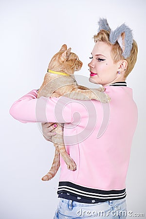 Nice kind woman with a short haircut and fur ears in sports clothes and jeans is holding her beloved pet on a white background in Stock Photo