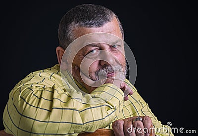 Portrait of a smiling senior man in yellow shirt leant elbow on a back of chair against black Stock Photo