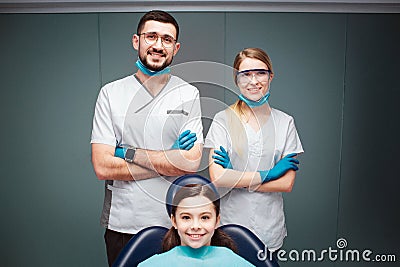 Nice positive male and female dentist with girl in dental chair. They look straight and smile. Adults hold hands crossed Stock Photo