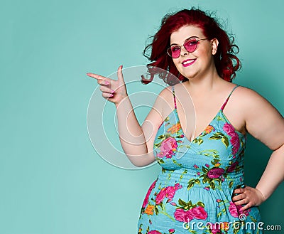 Nice smiling to us plus-size overweight lady in sunglasses and sundress point her finger at something on text copy space on mint Stock Photo