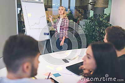 Nice young blonde woman look on camerea. She stand at flipchart and draw grafic line. Another people have conversation Stock Photo