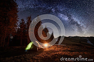 Night camping. Illuminated tent and campfire near forest under night sky full of stars and milky way Stock Photo