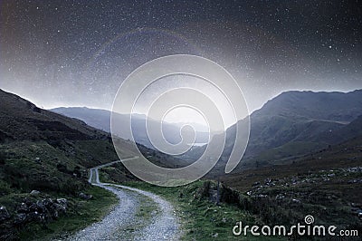 A night concept edit. A mountain track heading into the distance at night with stars in the sky with a bright light glowing on the Stock Photo