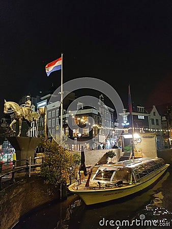 Night image of Amsterdam. Boat moored next to the flag of Holland and a statue of a man and a horse. Chain of lamps immunize the Editorial Stock Photo
