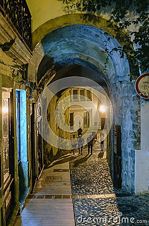 Night shot of a tunnel in a narrow, lonely alley with cobblestone flooring with unrecognizable people strolling and glowing from t Editorial Stock Photo