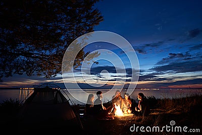 Night summer camping on shore. Group of young tourists around campfire near tent under evening sky Stock Photo