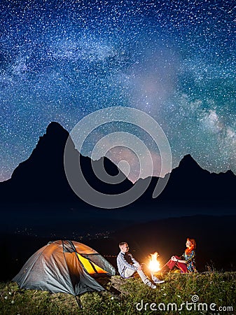 Night tent camping. Young couple backpackers sitting by bonfire under incredibly beautiful starry sky and Milky way Stock Photo