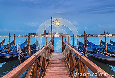 Evening Venice seascape with gondolas at moorings, light of lantern at wooden bridge, view at famous San Giorgio Maggiore island Stock Photo