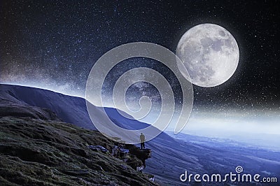 A nightime edit. A hiker on the edge of a cliff surrounded by mountains with the moon and stars above Stock Photo