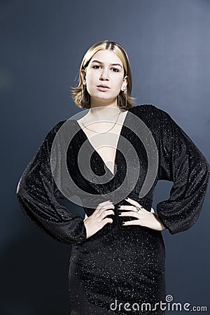 A nineteen-year-old girl with bleached strands and a bob hairstyle in a fashionable black glitter cocktail dress smiles. The dark Stock Photo