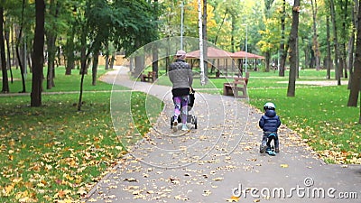 Niño pequeño montando moto balanceada moto por pasarela asfaltada junto con madre caminando cerca del camino del parque de la ciu almacen de video Niño pequeño montando moto balanceada moto por pasarela asfaltada junto con madre caminando cerca del camino del parque de la ciu almacen de video