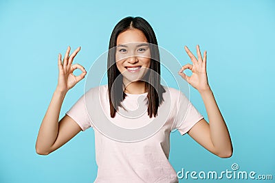 No problem. Smiling asian woman showing okay signs, approval gesture, pleased and satisfied, standing over blue Stock Photo