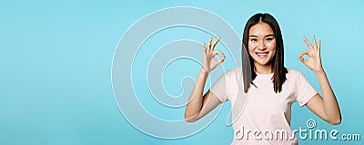 No problem. Smiling asian woman showing okay signs, approval gesture, pleased and satisfied, standing over blue Stock Photo