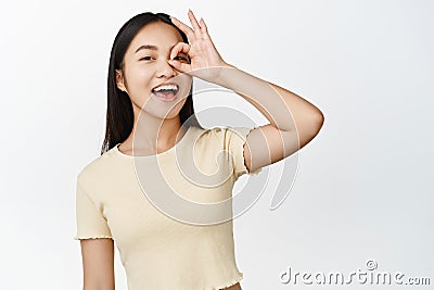 No problem. Smiling happy asian woman showing zero, okay gesture on eye, laughing carefree, standing over white Stock Photo