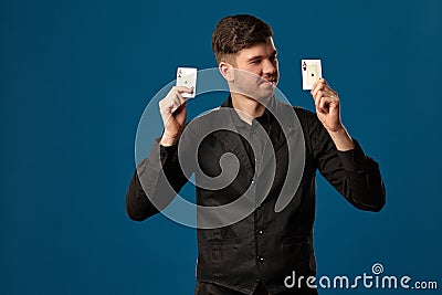 Noob in poker, in black vest and shirt. Holding two playing cards while posing against blue studio background. Gambling Stock Photo