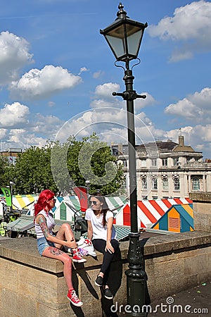 NORWICH, UK - JUNE 3, 2017: Two British young women enjoying a sunny day at Memorial Gardens with the colorful stalls of an open-a Editorial Stock Photo