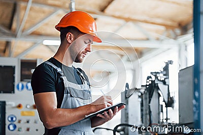 With notepad in hands. Industrial worker indoors in factory. Young technician with orange hard hat Stock Photo