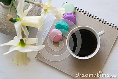 Notepad, macarons, cup of coffee and daffodils in a vase on a white table. Inspirational workplace. Stock Photo