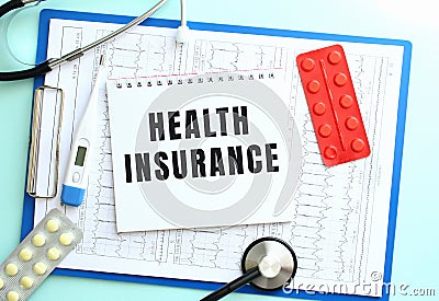 A notepad with the text HEALTH INSURANCE lies on a medical clipboard with a stethoscope and pills on a blue background. Stock Photo