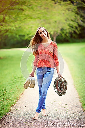 Nothing can steal my smile today. a young woman walking in a park holding her shoes and a hat. Stock Photo