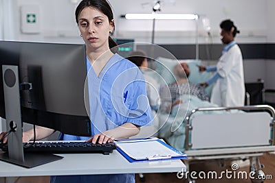 Nurse at medical desk typing patient data into computer while team of doctors putting iv drip line Stock Photo