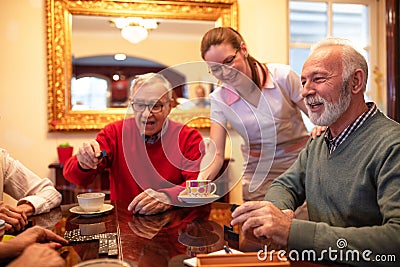 Nurse serving tea while older people play board games, retirement home Stock Photo