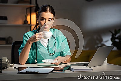 Nurse in uniform sitting at table and drinking coffee during night shift Stock Photo