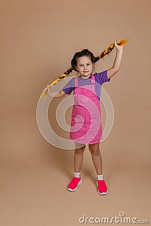 Obedient calm little child hodling yellow kanekalon pigtails looking at camera with slight smile in pink jumpsuit and Stock Photo
