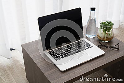 office table with blank screen on laptop, bottle of water, modern glasses, garden plant on glass vase on white drape background t Stock Photo