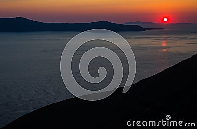 Oia santorini island greece. Natural coloured sunset. Dark red sky. Gold setting sun behind mountains. Calm sea reflecting Stock Photo