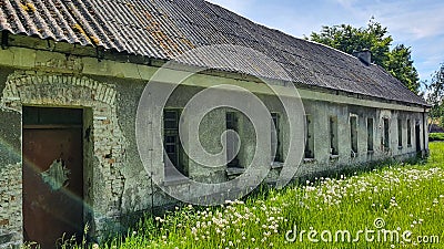 Old abandoned long house in poor condition. Bars and cracked panes in the windows. Around the tree and dandelions in the grass. Stock Photo