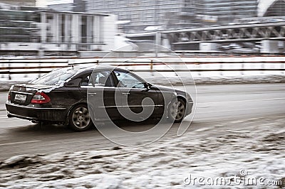 Old black Mercedes W211 on snowy winter road in city. Mercedes-Benz E-class drives on slippery snow covered street Editorial Stock Photo