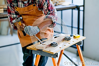 Old craftsman use hammer and chisel to work with wood and produce some product in his workplace during day time Stock Photo