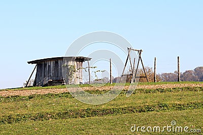 Old dilapidated wooden tool shed with cracked walls next to large wooden frame swing on top of small hill Stock Photo