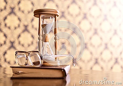 Old fashioned attributes concept. Hourglass, old book and eyeglasses on wooden table, pattern background. Hourglass Stock Photo
