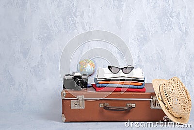 Old fashioned travel suitcase with summer accessories: glasses, pack of clothing, retro photo camera, straw beach hat on grey back Stock Photo