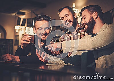 Old friends having fun with smartphone and drinking draft beer at bar counter in pub. Stock Photo