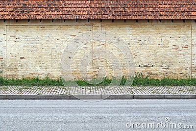 Old grunge yellow brick wall with terracotta tiled roof. A stripe of grass, concrete tiled sidewalk and asphalt road in front. Stock Photo