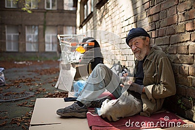 Old homeless man sitting on cardboard holding bowl with food in hand. Stock Photo