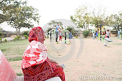 An old indian lady sitting on the bench and watching children playing in an open gym in a park Editorial Stock Photo